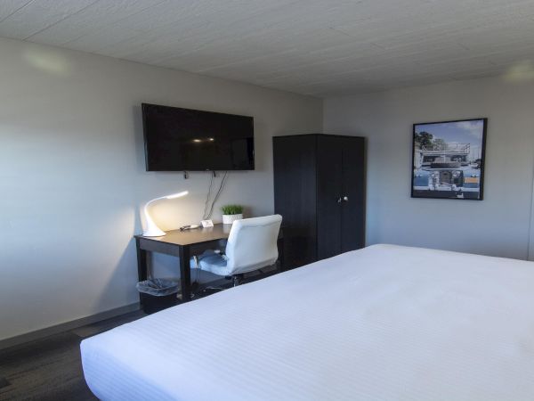 A modern hotel room with a large white bed, a small desk and chair, a flat-screen TV, a wardrobe, and a framed photo on the wall.