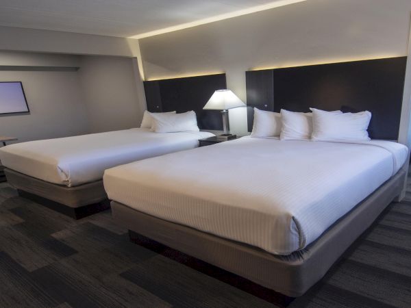 A modern hotel room with two large doubles, plush beds, nightstands, lamps, a wall-mounted TV, and a desk area; clean and contemporary.