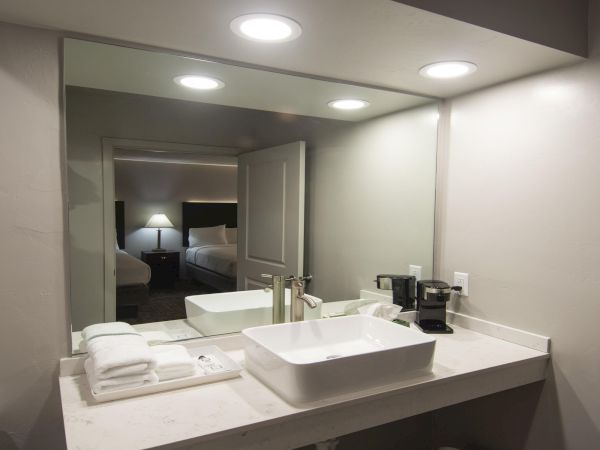 Hotel bathroom with a marble counter, square vessel sink, towels, and a large mirror reflecting a bedroom with bed and nightstand, all under recessed lighting.