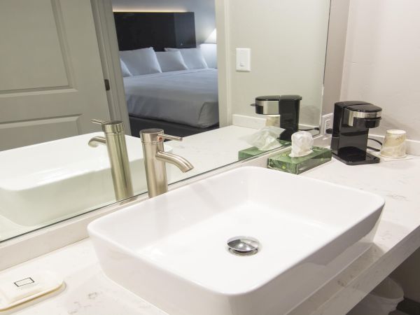 A modern hotel bathroom with a double sink, marble counter, wall mirror, and a coffee station on the counter near a doorway to a bedroom.