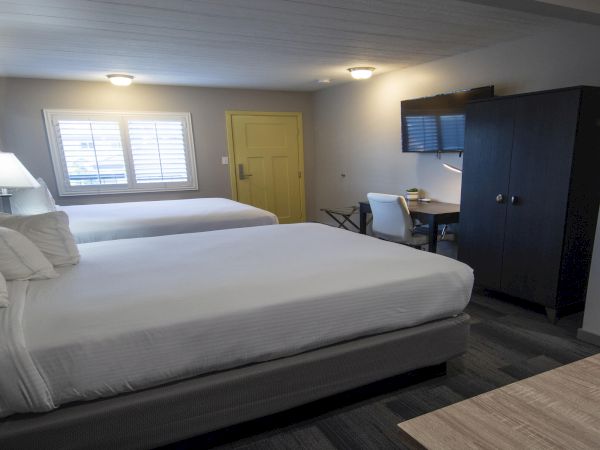 Two double beds in a hotel room with a yellow door, nightstand lamps, a desk and chairs, a wardrobe, and a small coffee/coffee station area, all lit softly.