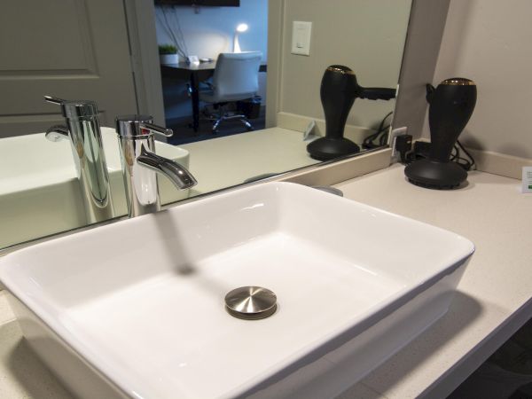 A clean bathroom vanity with a rectangular white sink, chrome faucet, large mirror, two black hairdryers, and a small card on the counter.