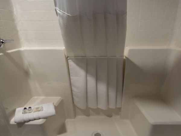 A compact, white shower stall with built-in benches on both sides, a fold-down curtain, and a small soap/towel on a wall shelf, ending abruptly.
