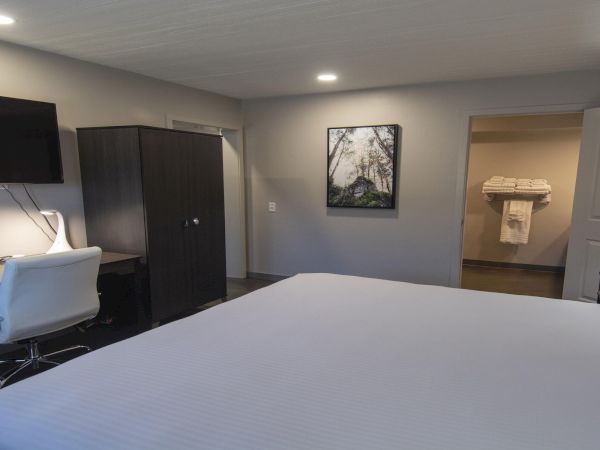A tidy hotel room with a large bed, a desk and chair, a wall-mounted TV, a dark wardrobe, and a small seating area; after-door to a bathroom with towels.