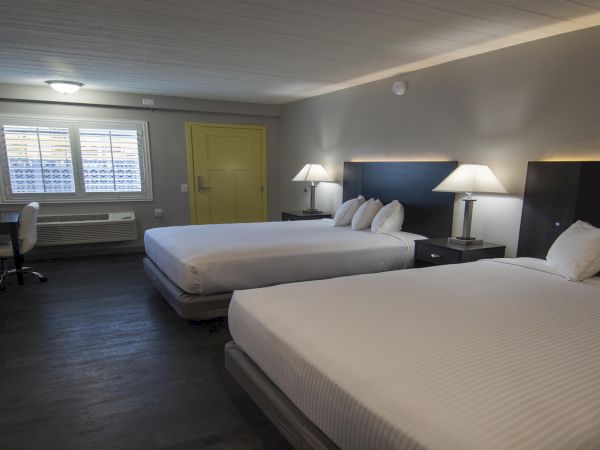 A hotel room with two beds, lamps, a desk and chair, window blinds, and a green door.