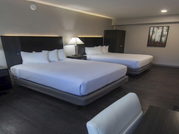 A modern hotel room with two large beds, two nightstands, lamps, a small desk, a cabinet, and a framed picture on the wall.