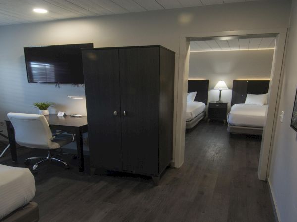 A modern hotel room with two beds in the back, a desk and chair, a dark wardrobe, and a separate seating area, all in neutral tones.