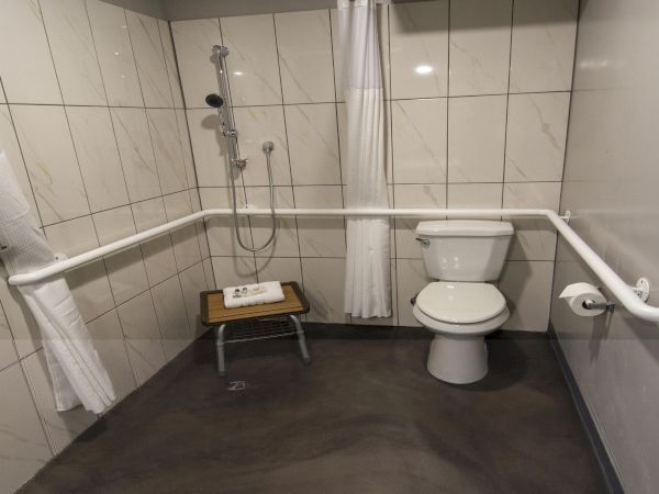 A male or female accessible bathroom with a toilet, a handheld shower, grab bars, a small stool, and towel shelves in a tiled room.