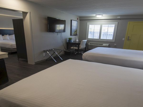 Two neatly made double beds in a bright hotel room with a yellow door, mounted TV, desk, mini fridge, and a small seating area by the window.