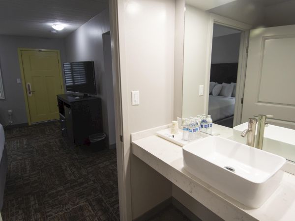 A hotel bathroom and entry area with a modern sink on a counter, a mirror, and a view into a dimly lit room with a yellow door and TV, ending with a period.