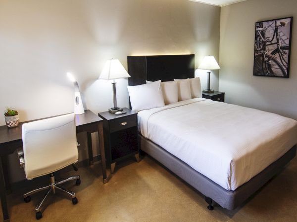 A hotel room with a large bed, two nightstands and lamps, a desk with a chair, a plant on the desk, and wall art, all neatly arranged.