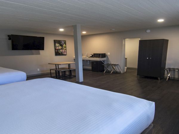 A spacious hotel room with two large beds in the foreground, a desk and chair, kitchenette area, flat-screen TV on the wall, and a dark wardrobe with a small table and stools.