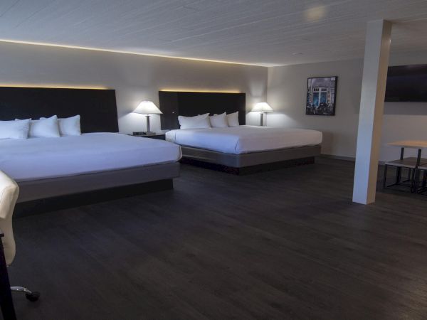 Two large hotel rooms with two queen beds each, sleek dark wood floors, nightstands, lamps, a desk chair, and a small dining table in a bright, modern suite.