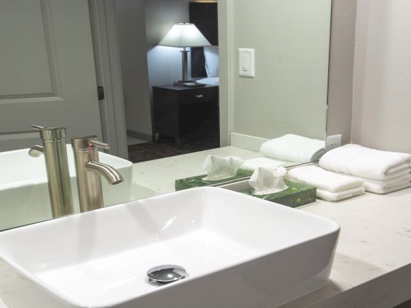 A bathroom countertop with a rectangular sink, faucet, folded towels, and a mirror reflecting a lamp and bedroom in the background, nice and tidy.