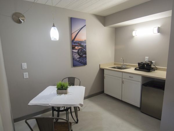 A small kitchenette with a table for two, pendant light, plant centerpiece, mini-fridge, sink, and a coffee maker on the counter, compact and modern.
