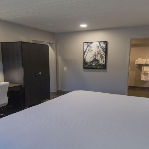 A clean hotel room with a large bed, desk and chair, mounted TV, wardrobe, and a view into a bathroom area with towel rack; neutral tones.