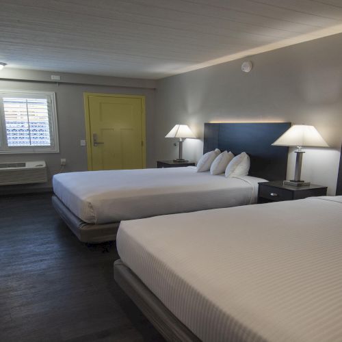 A modern hotel room with two beds, bedside tables and lamps, a desk, and a window with blinds.