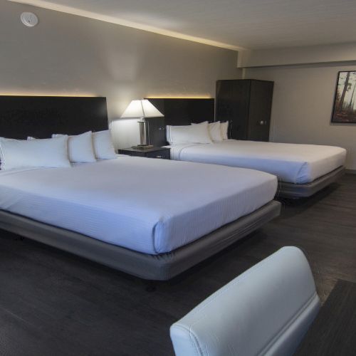 A modern hotel room with two king beds, bedside lamps, a desk, and framed wall art under bright ceiling lights.