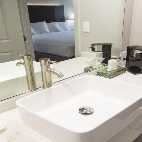 A modern hotel bathroom with a white countertop, vessel sink, chrome faucet, mirror, coffee station, and a view into a bedroom.
