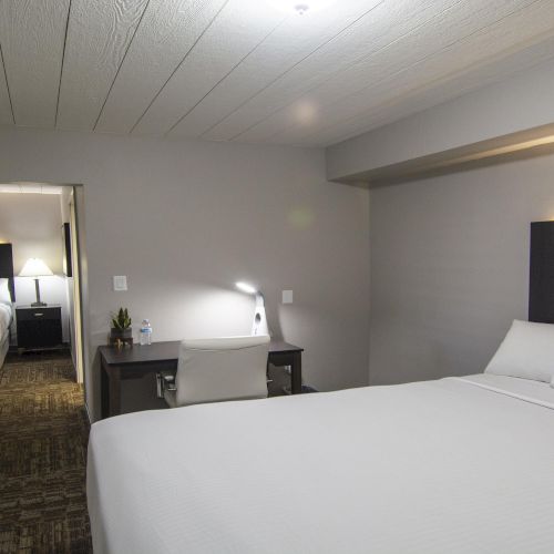 A modern hotel room with white bedding, two beds, a small desk, and a vanity area, lit by warm overhead lights.