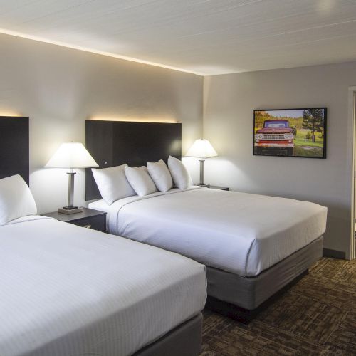 A bright hotel room with two beds, bedside lamps, framed wall art, a TV, and an open doorway.