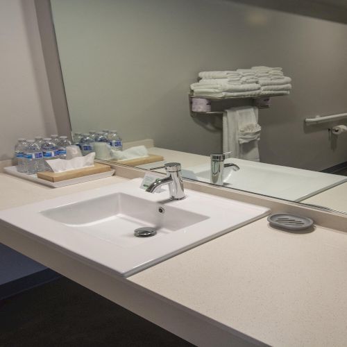 A clean hotel bathroom sink area with a double sink, mirror, stacked towels, water bottles, and a dark barware setup on the counter.