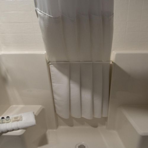 A compact shower stall with built-in seats, a small fold-down shelf, a handheld shower head, and a curtain in the center. The space is tiled and compact, with a drain in the floor.