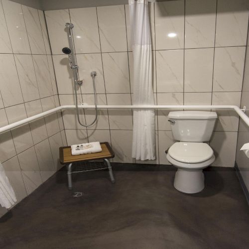 A clean modern bathroom with a shower, chrome fixtures, a toilet, and folded towels on a small wooden stool.