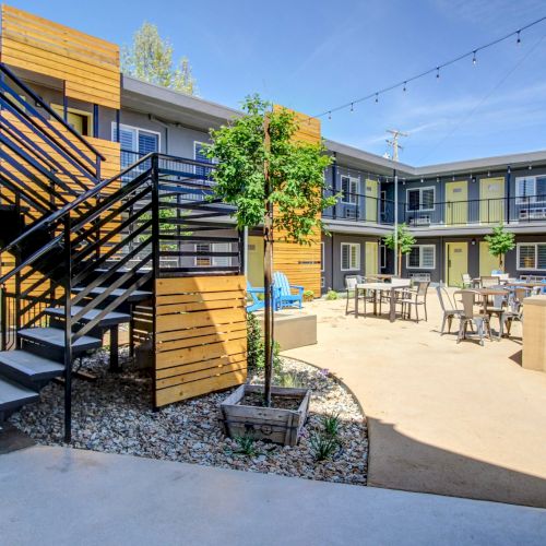 Sunny apartment courtyard with steps, patio tables, string lights, and greenery under a clear blue sky.