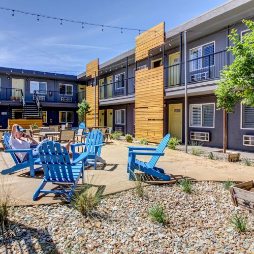 Courtyard of a modern apartment complex with blue lounge chairs, a man relaxing, string lights, and trees against sunny sky.