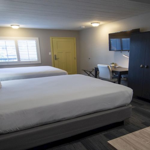 A modern hotel room with twin beds, bedside lamps, a desk with chair, a flat-screen TV, and gray cabinetry.