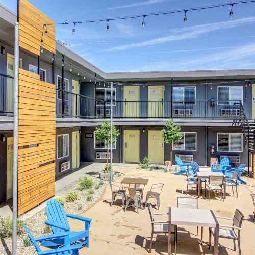 A sunny apartment courtyard with yellow doors, blue chairs and tables, and hanging string lights between buildings.