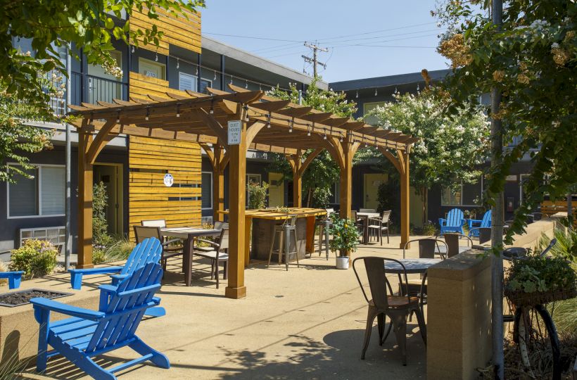 An outdoor patio with wooden pergolas, tables, and colorful chairs (blue, brown, and black) in a sunny courtyard, likely a cafe or resort.