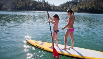 Two kids paddle a yellow SUP on a lake, standing and using a red paddle near forested mountains and clouds.
