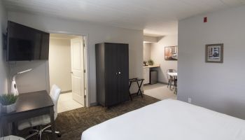 A modern hotel room: bed, desk with chair, wall TV, dark wardrobe, and a small sitting area in the back.