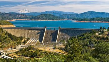 A large concrete hydroelectric dam controls a turquoise lake, with forested hills and snowy mountains in the background.
