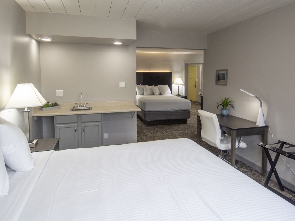 A modern hotel room showing two beds, a small kitchenette with sink, a desk and chair, and a TV-lit wall.