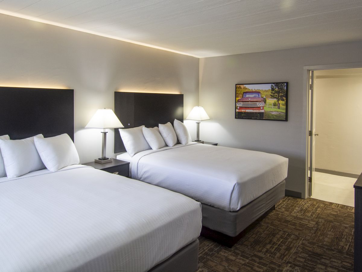 A hotel room shows two queen beds, bedside lamps, framed car artwork, a TV, and an open door.