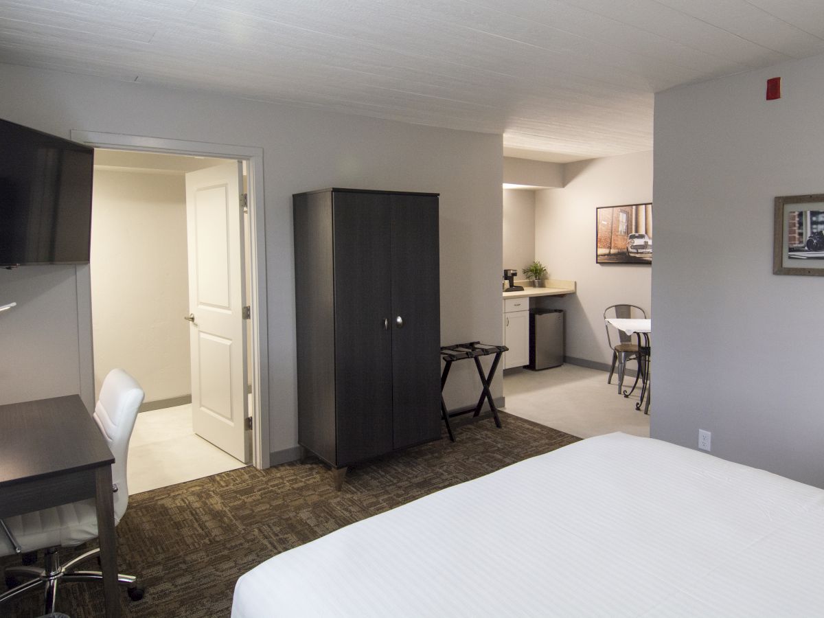 A modern hotel room with a bed, desk, mounted TV, dark wardrobe, and a small kitchenette area in the background.