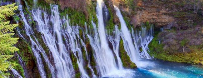 A wide cascade of waterfalls pours over a rocky, mossy hillside into a bright blue river in a forested canyon, .