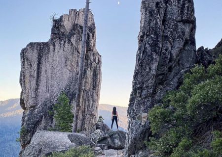 A hiker stands on a rocky mountain ledge, silhouetted between two towering stone cliffs under a clear blue sky.
