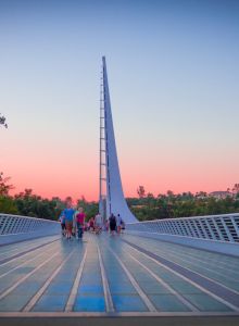 A modern pedestrian bridge with a tall central spire and blue-tinted glass walkway, sunset sky, and people strolling outward.