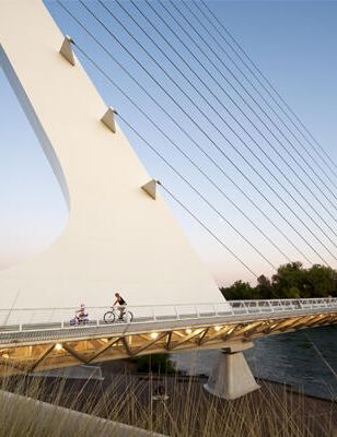 A cyclist rides across a modern cable-stayed pedestrian bridge over a river under a clear blue sky.