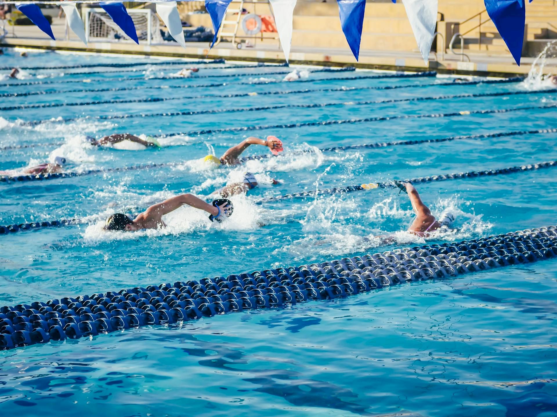 Swimmers race in a blue indoor pool during a meet, splashing between lane ropes under blue-and-white flags.
