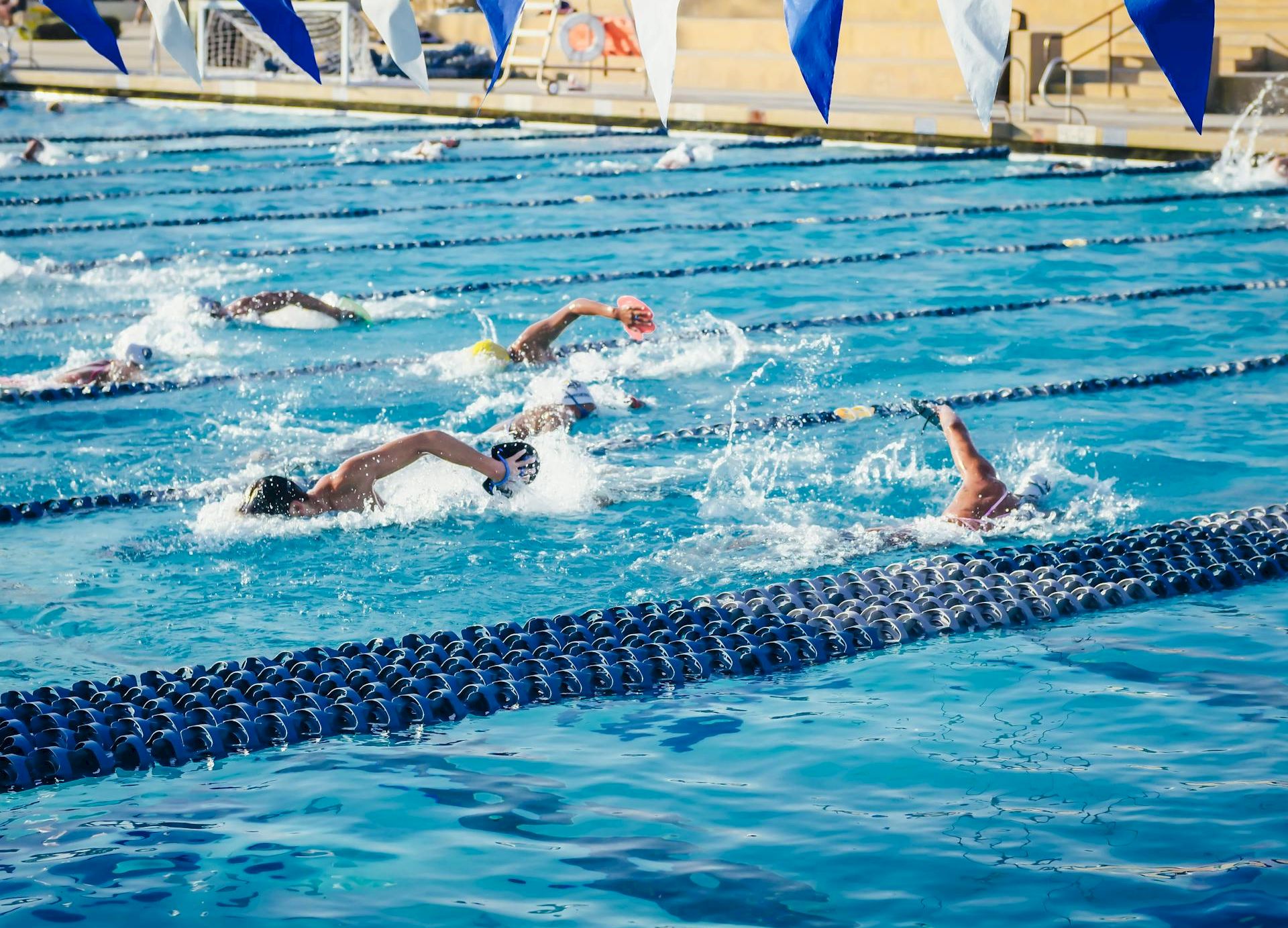 Swimmers race in a blue indoor pool during a meet, splashing between lane ropes under blue-and-white flags.