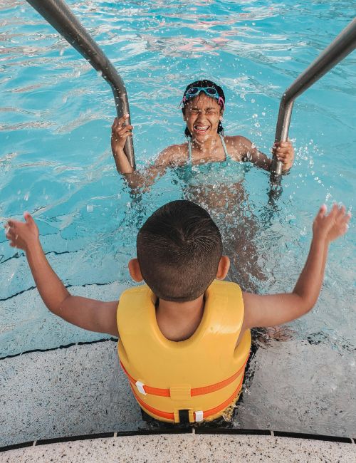A smiling person in a pool, wearing swim goggles and holding the boat&rsquo;s rails while splashing water around them, with a child in an inflatable float below, having fun!