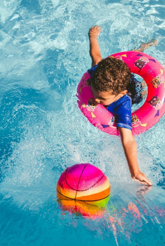 A child swims in bright blue water with a pink float ring and rainbow pool ball in a sunny splash.