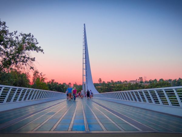People stroll on a modern bridge at sunset, with a towering spire rising ahead into a pink-blue sky.