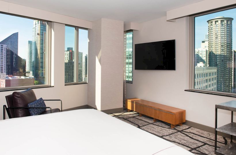 Modern hotel room with large bed, wall TV, and floor-to-ceiling windows overlooking a city skyline in daylight.