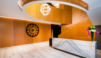 Modern hotel reception desk in a wood-paneled lobby with a large wall clock, globe light, and a flower vase.